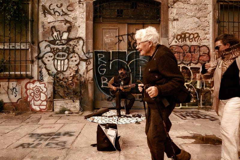 A smiling white-haired woman holding a walking pole passes a guitarist who sings into a microphone in front of a graffiti-covered doorway, as another woman in sunglasses walks in from the right.