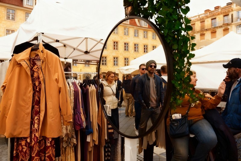 An oval market mirror reflects pedestrians beside hanging clothes and two people in conversation.