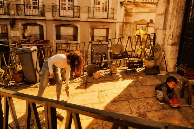 An adult bends down while a child plays beside plant pots on a sunlit stone landing.